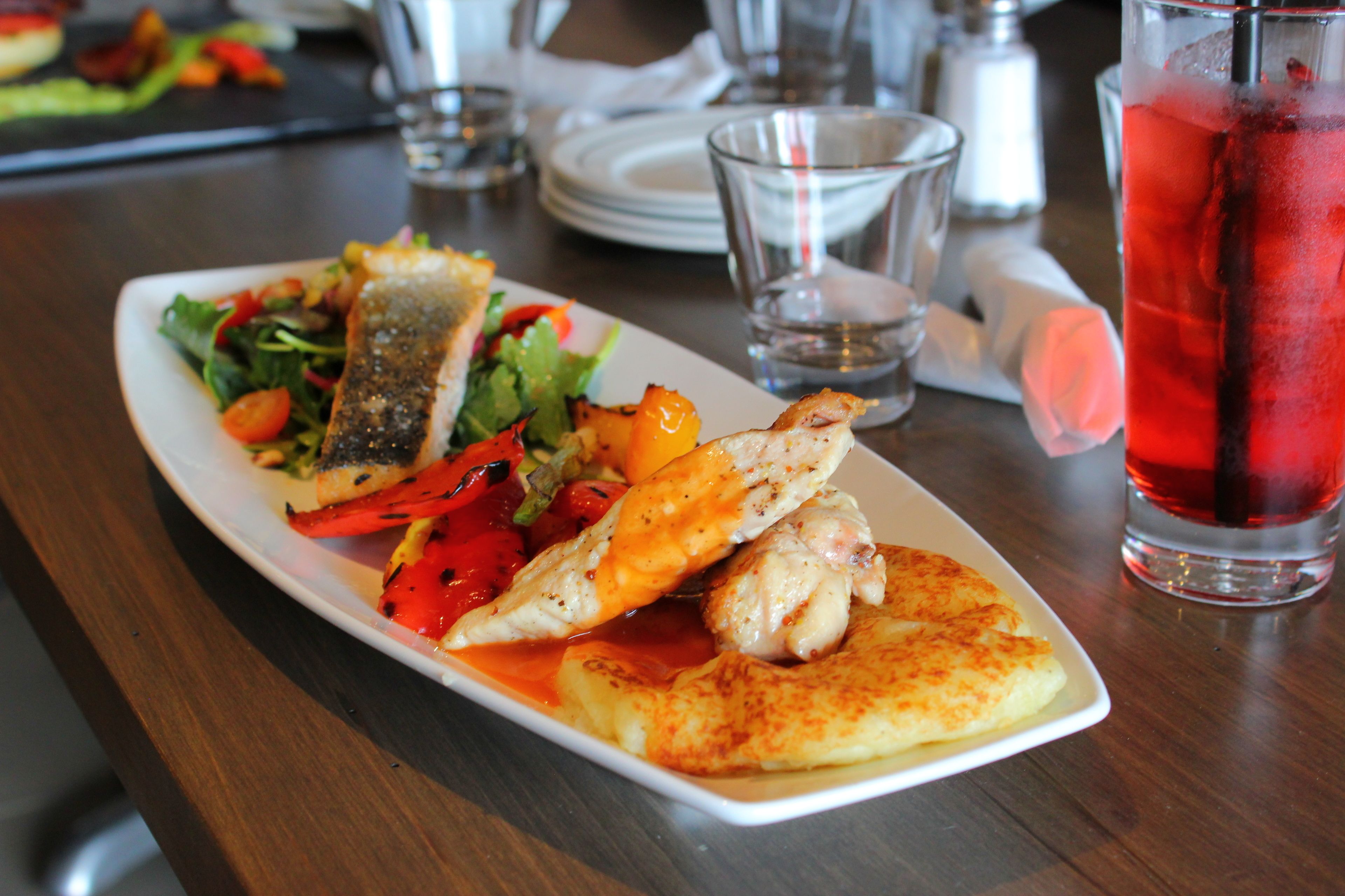 A plate of food with grilled vegetables and fish, a glass of red drink, and a white napkin on a wooden table.