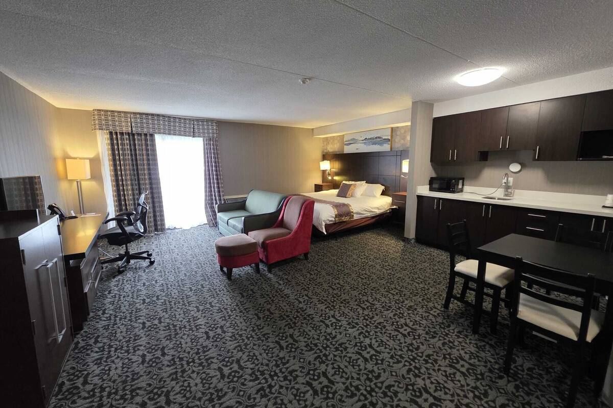 A hotel room with a bed, a desk, a chair, a sofa, a dining table, and a kitchenette.