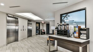 Un área de ascensores moderna con ascensores de acero inoxidable, un mostrador de recepción con máquinas de café y un amplio ventanal con vistas.