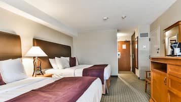 A hotel room with two beds, a wooden dresser, a lamp, and a hallway leading to a door.