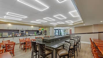 Un área de cocina moderna con una isla central, taburetes de bar y una zona de estar con mesas y sillas de madera.