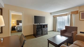 Habitación de hotel con televisión de pantalla plana, minibar, zona de estar con mesa y sillas, y un ventanal con cortinas.