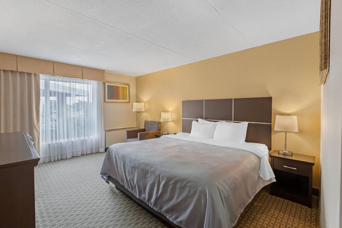 A hotel room with a large bed, two bedside tables with lamps, a desk, and a window with sheer curtains.