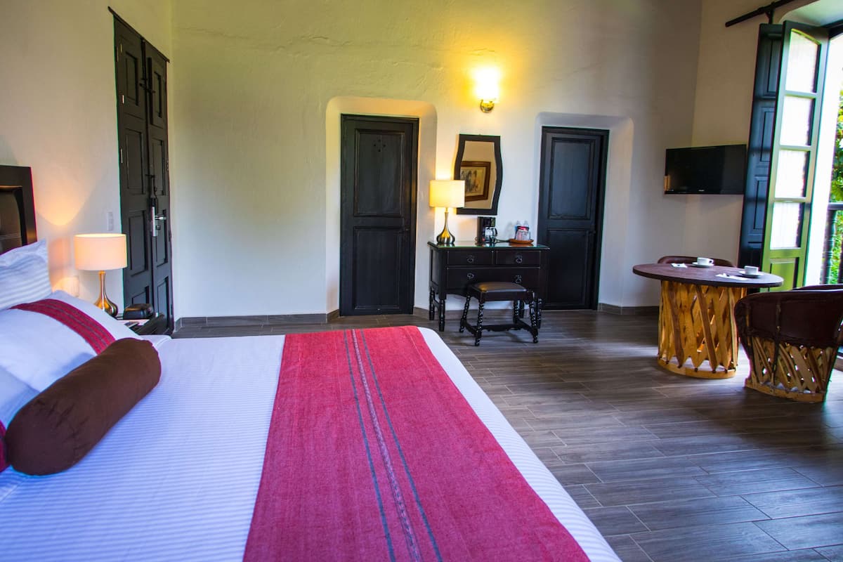 Ein Hotelzimmer mit einem großen Bett, einem Schreibtisch mit Lampe, einem Sessel, einem kleinen runden Tisch und einem an der Wand befestigten Fernseher.