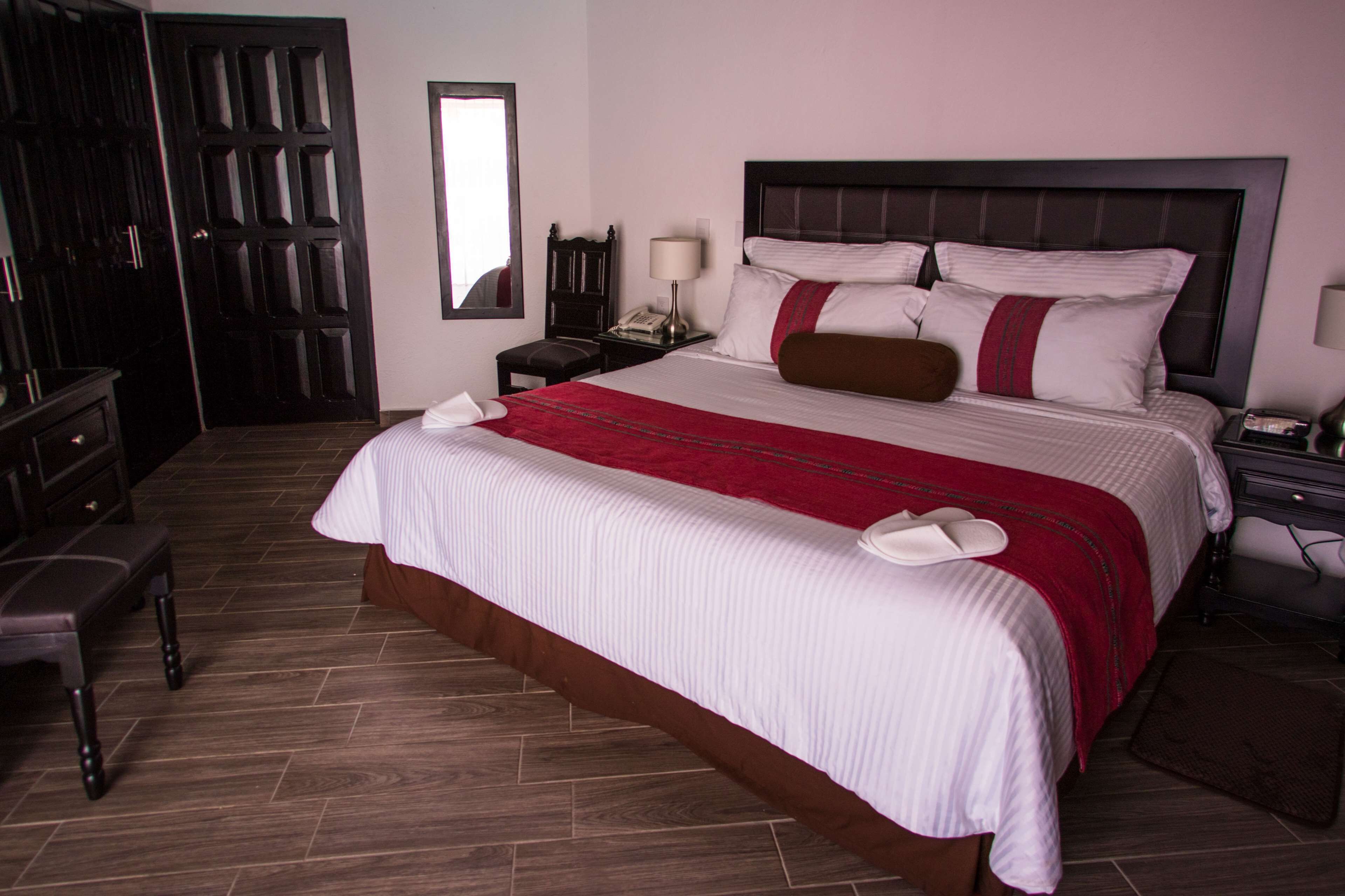 A neatly made bed with a red and white bedspread, a wooden nightstand with a lamp, and a dark wooden wardrobe.