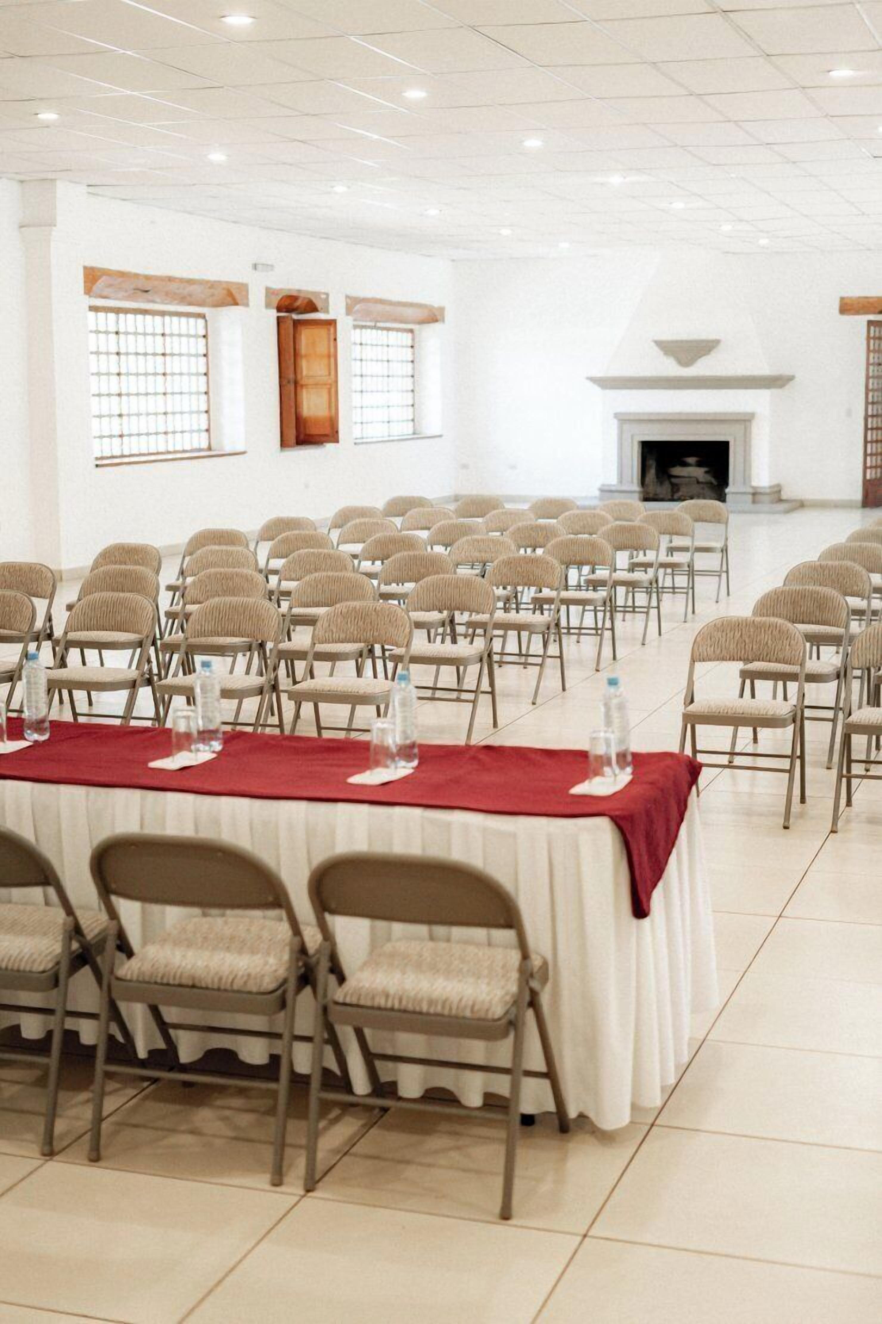 A conference room with rows of chairs, a long table with a red tablecloth, and a fireplace in the background.