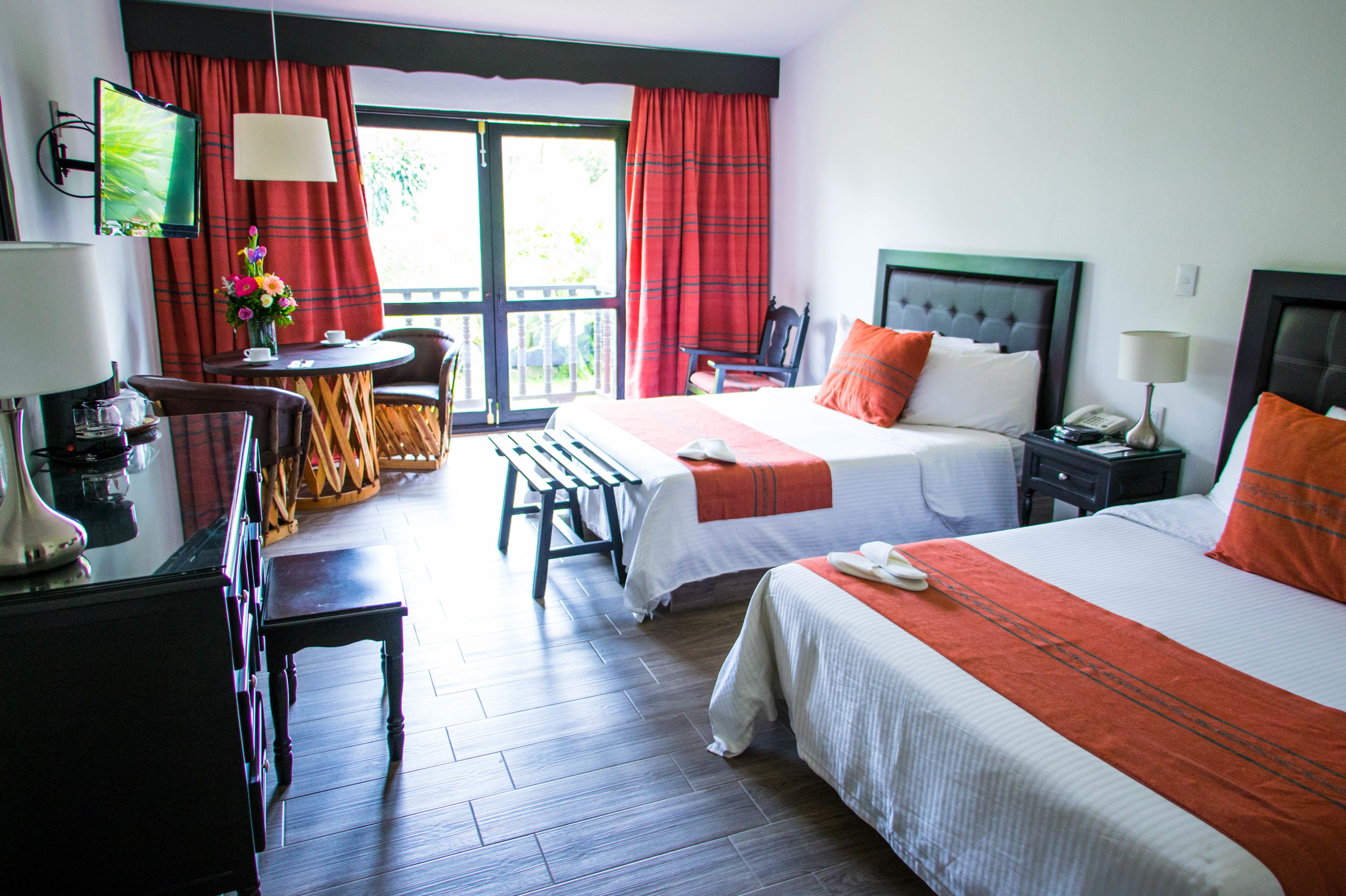 A hotel room with two beds, a desk with a chair, a TV, and a balcony with red curtains.