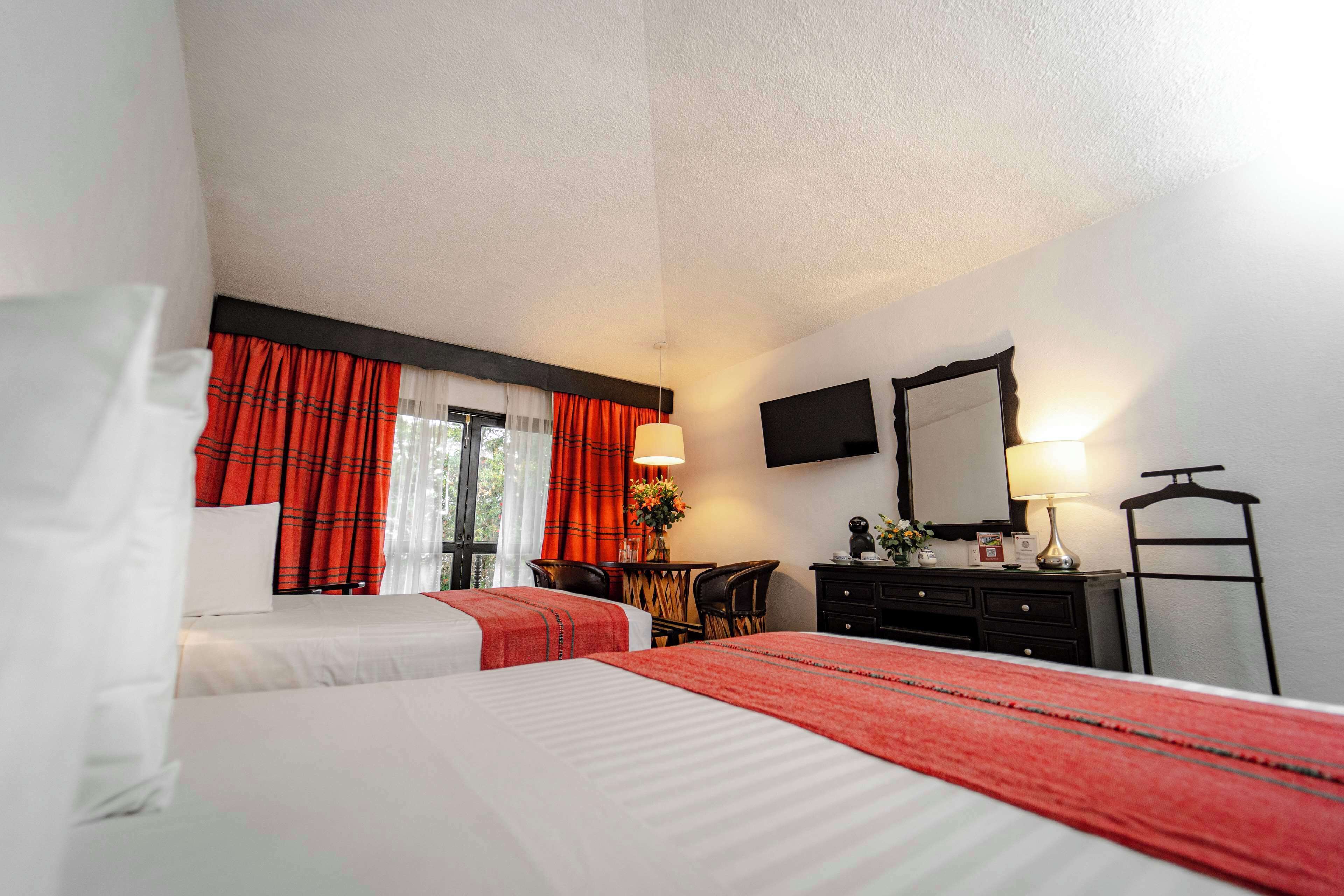 A hotel room with two beds, a desk, a chair, a dresser, a TV, and a mirror.