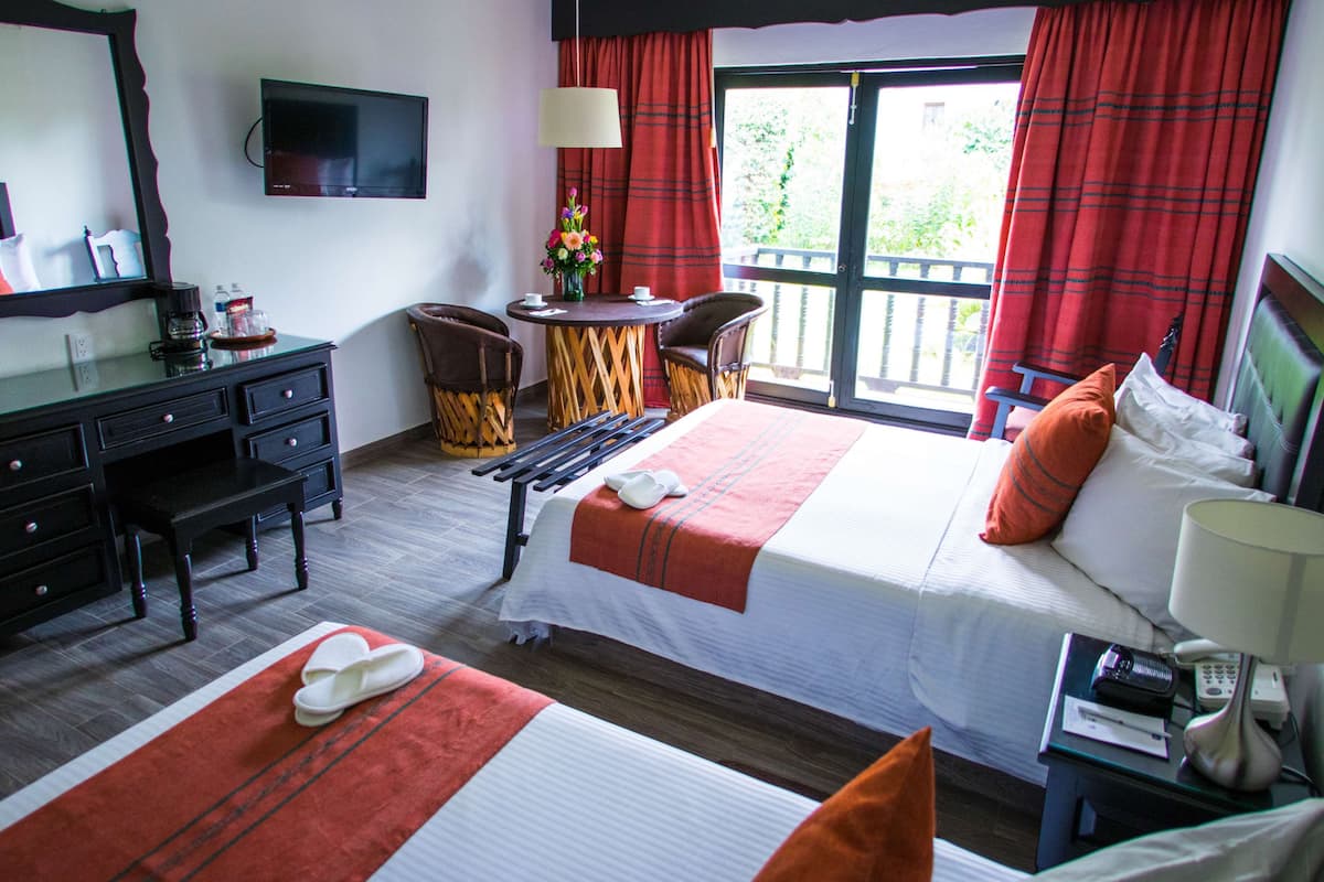 Ein Hotelzimmer mit zwei Betten, einem Fernseher, einem Schreibtisch mit Stuhl, einem kleinen Tisch mit Blumenvase und einem Balkon mit roten Vorhängen.