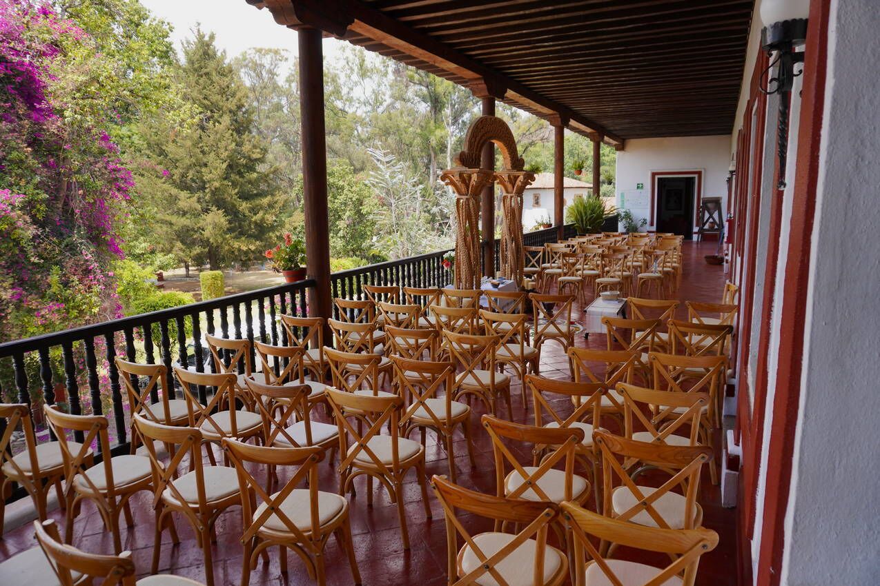 Un espace extérieur aménagé avec des meubles de jardin en bois, comprenant des chaises et des tables, et entouré d’une rambarde. Il offre une vue sur un jardin agrémenté d’arbres et de plantes en fleurs.
