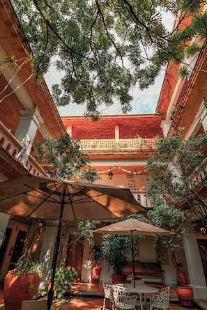 Une cour avec une table et des chaises placées sous un grand parasol, entourée de plantes en pot et d’un bâtiment en briques rouges.