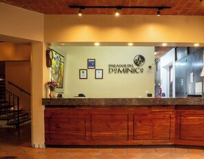 Un comptoir d’accueil avec un plan de travail en bois, une horloge, des certificats encadrés et une enseigne murale indiquant « PARADOR DEL DEMINICO ».