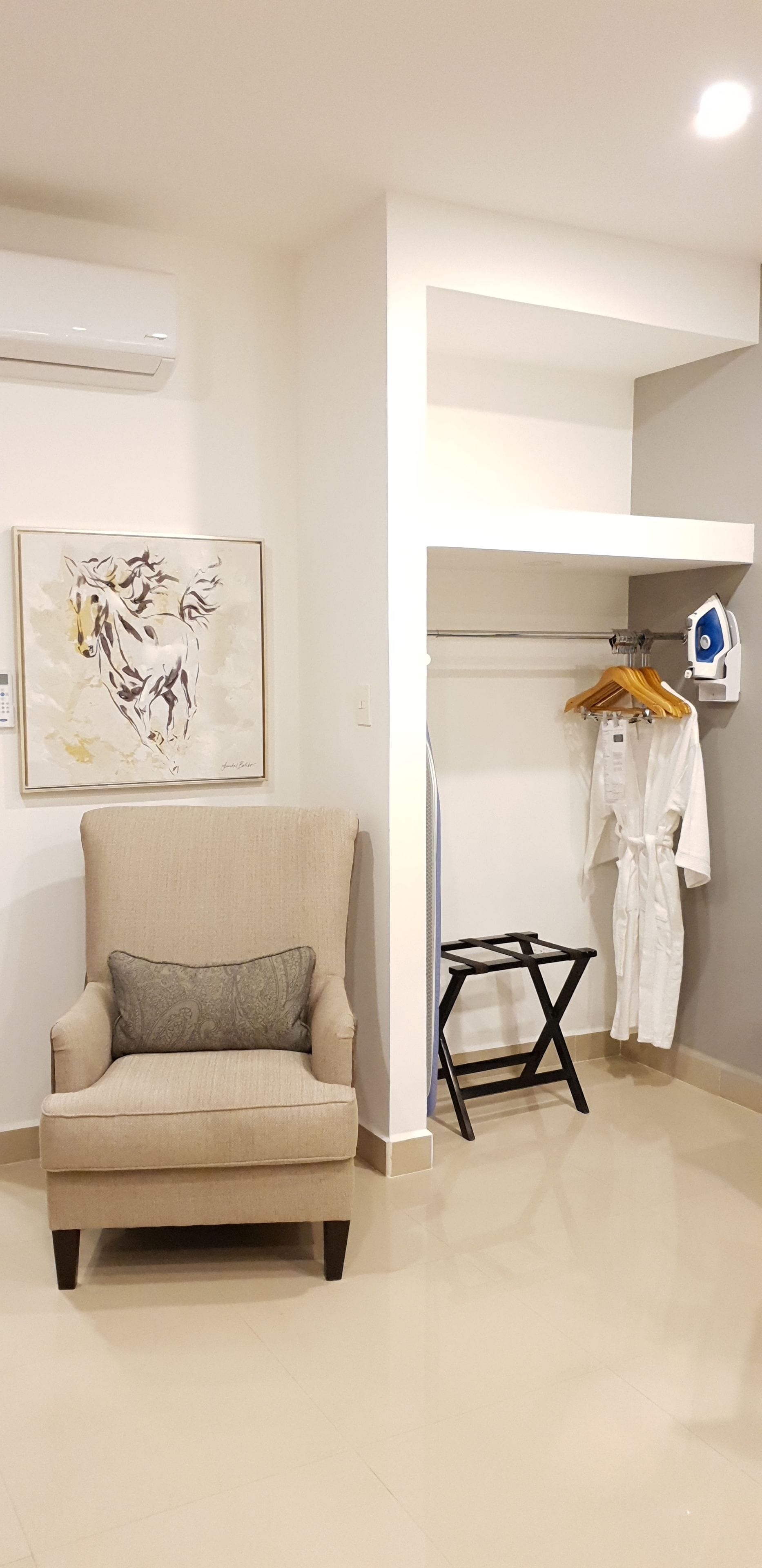 Une pièce comprenant un fauteuil beige, une petite table, une planche à repasser fixée au mur et un peignoir suspendu à un porte-vêtements.