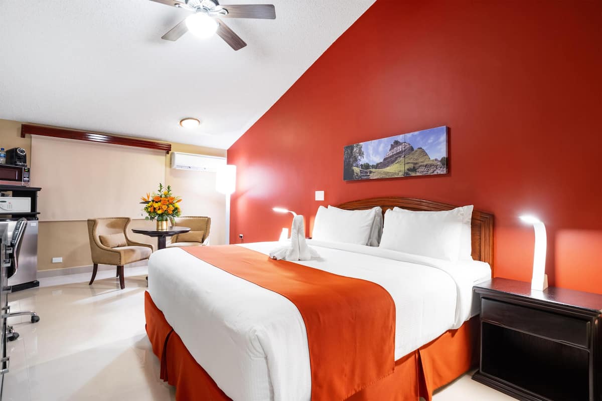 Habitación de hotel con una cama, un escritorio, una silla, una mesita con un jarrón de flores y un cuadro en la pared.