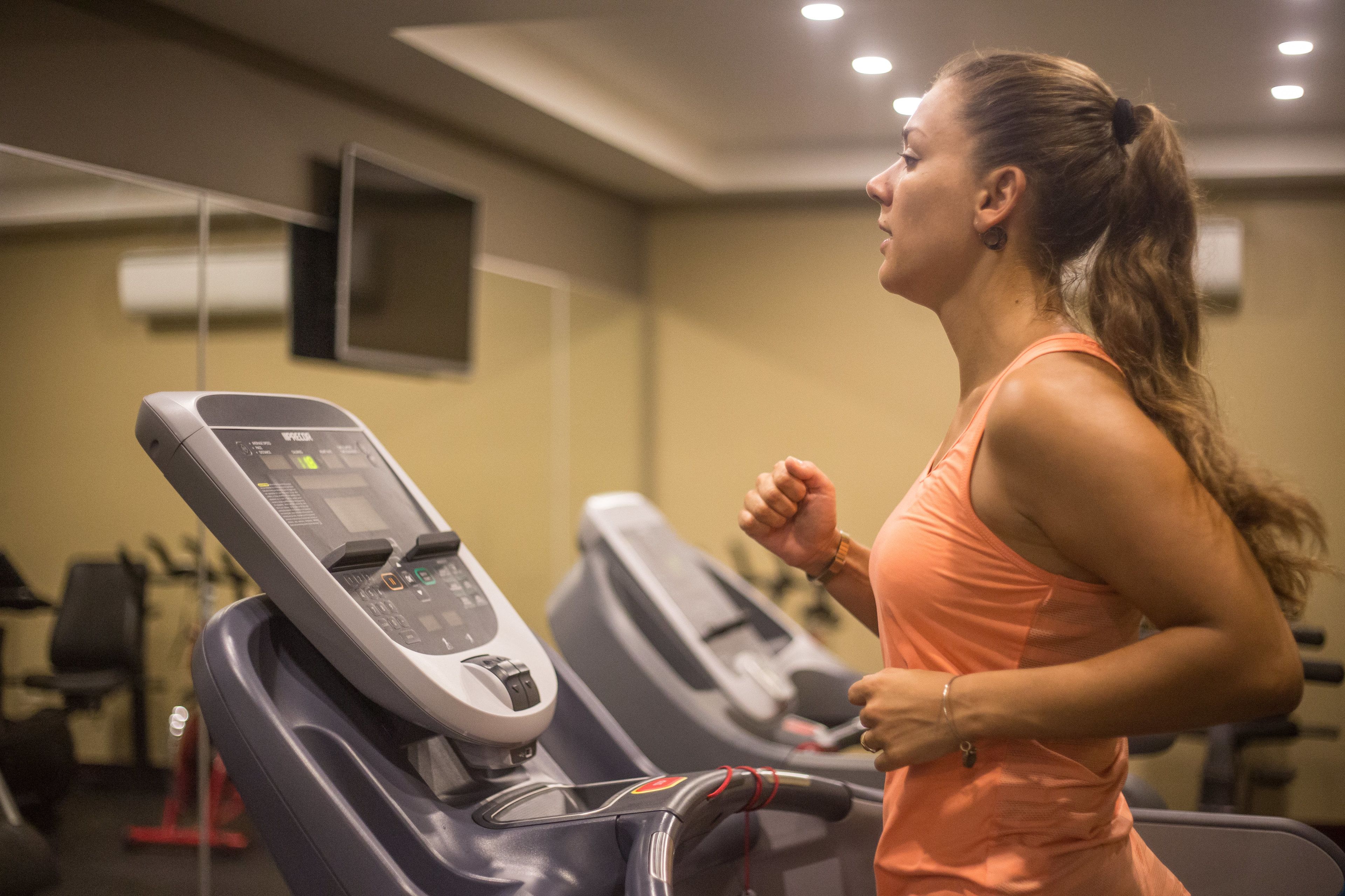 A woman running on a treadmill in a gym with a control panel and a television mounted on the wall.