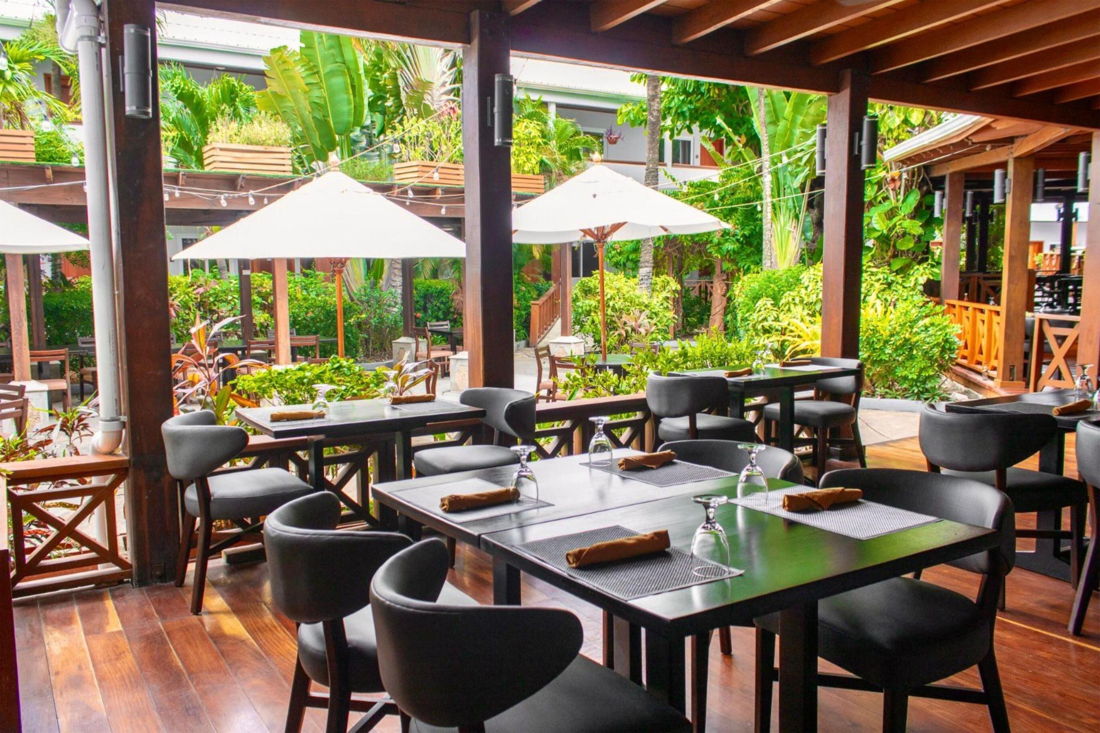 An outdoor dining area with tables and chairs, surrounded by lush greenery and covered by wooden pergolas.
