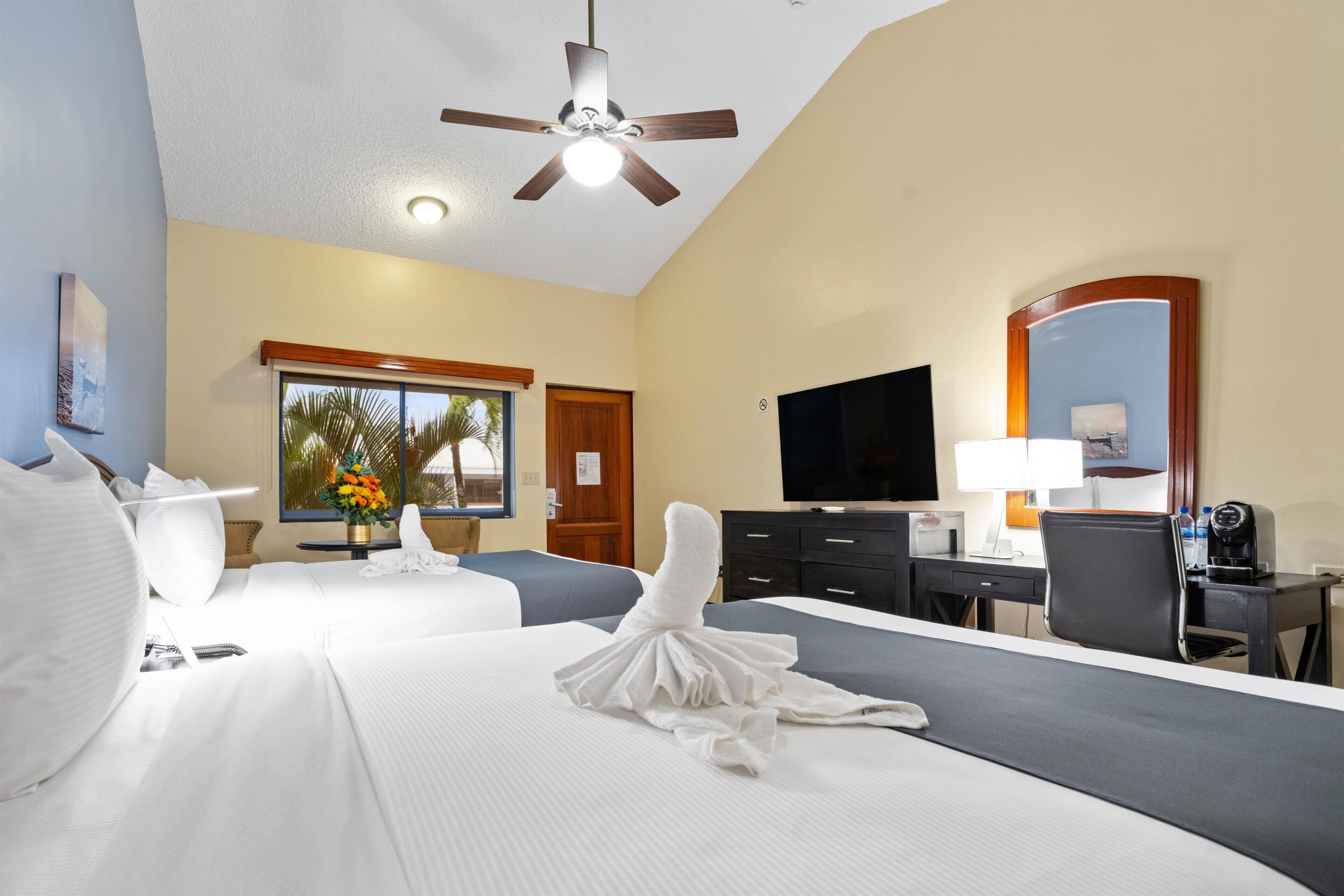 A hotel room with two beds, a desk, a chair, a television, a ceiling fan, and a window with a view of palm trees.