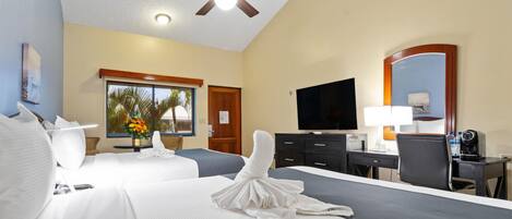 Habitación de hotel con dos camas, un escritorio, una silla, un televisor, un ventilador de techo y una ventana con vista a palmeras.
