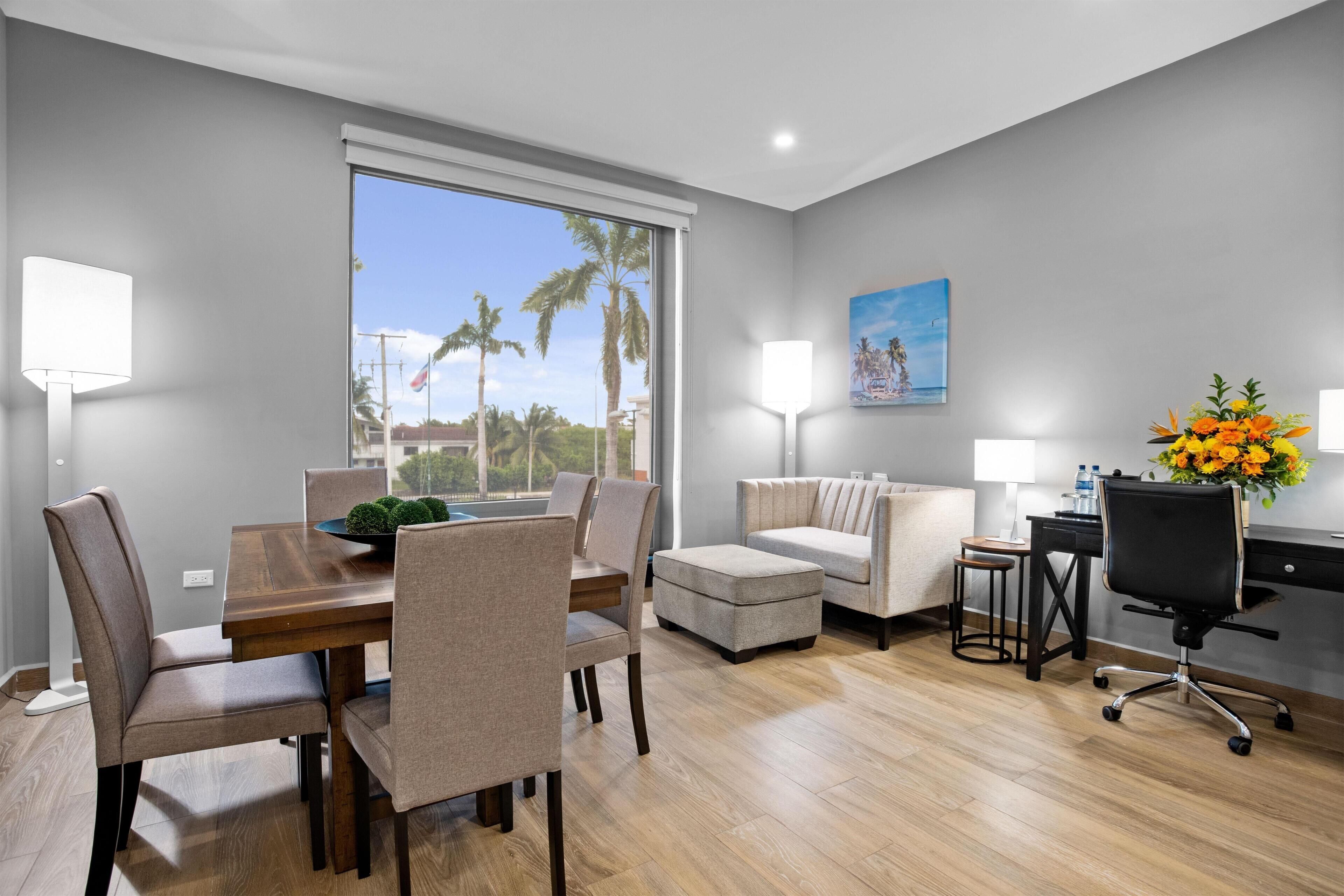 A modern hotel room with a dining area, a sofa, a desk, and a large window with a view of palm trees.