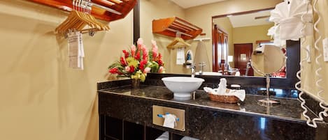 Un baño moderno con encimera de mármol negro, un lavabo blanco y un espejo grande. Se ven perchas de madera y un cesto con artículos de aseo en la encimera.