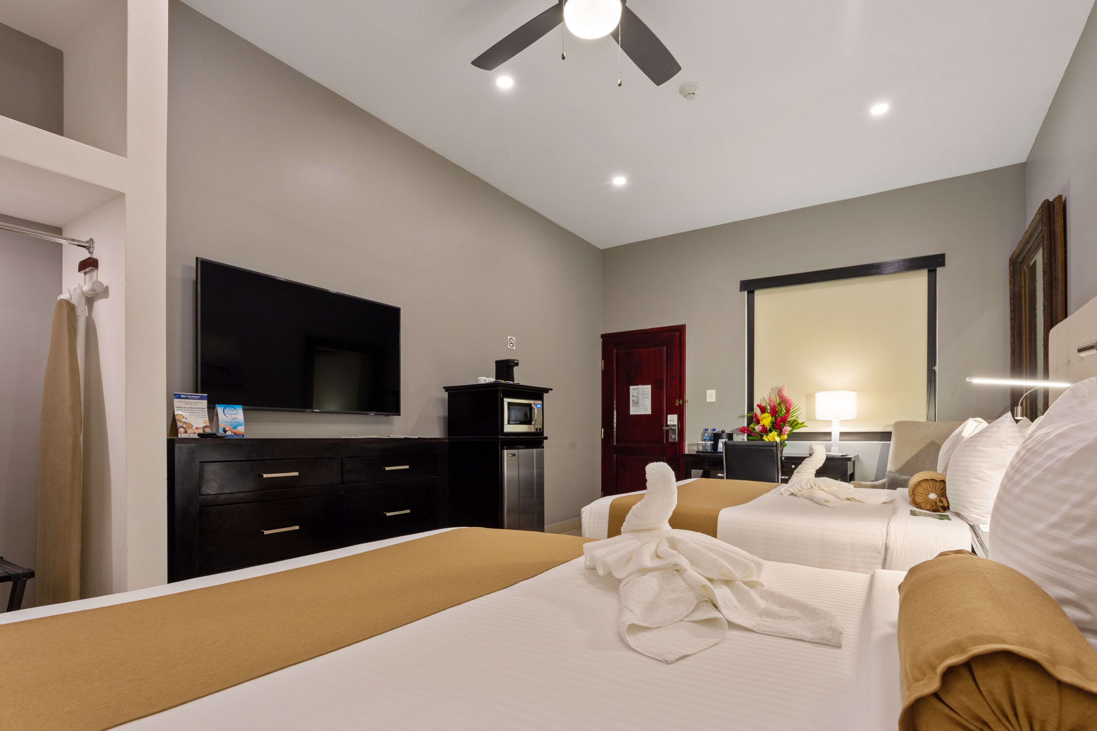 Une chambre d’hôtel avec deux lits, un téléviseur à écran plat, un bureau avec une lampe et un ventilateur de plafond.