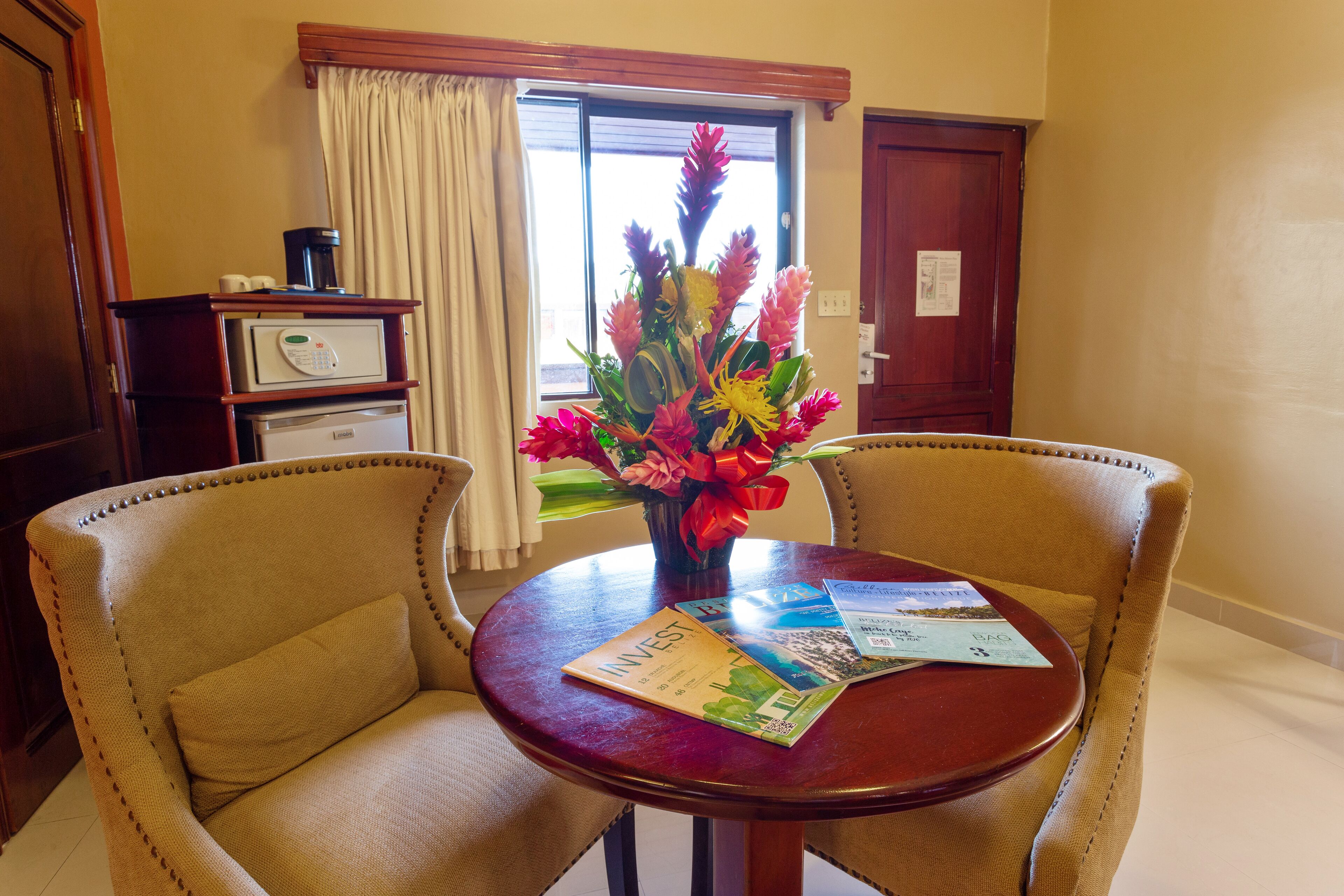 A hotel room with a round wooden table, two beige armchairs, a vase of flowers, a coffee maker, and a minibar.