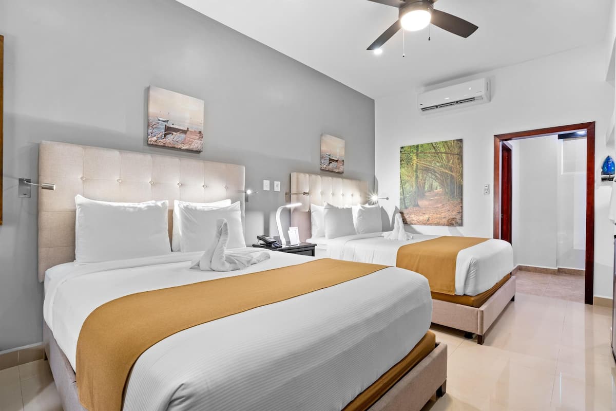 Habitación de hotel con dos camas, ventilador de techo, obras de arte en las paredes y una puerta que da a otra habitación.