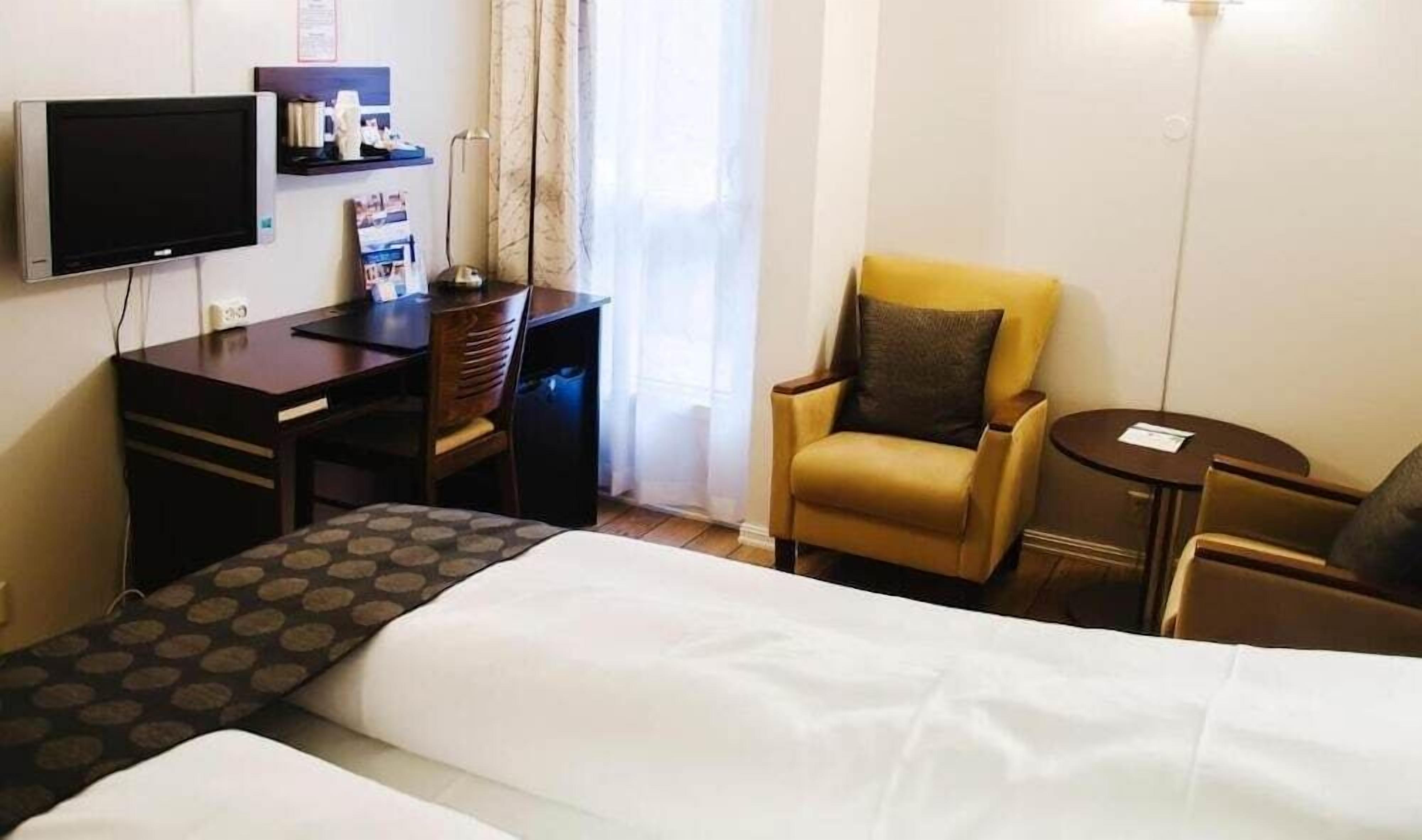 Une chambre d’hôtel avec un lit, un bureau avec une télévision, une chaise, une petite table et une fenêtre avec des rideaux.