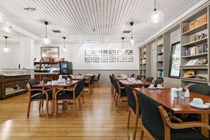 Un comedor con mesas y sillas de madera, una zona de cocina con una cafetera y una estantería con libros y objetos decorativos.