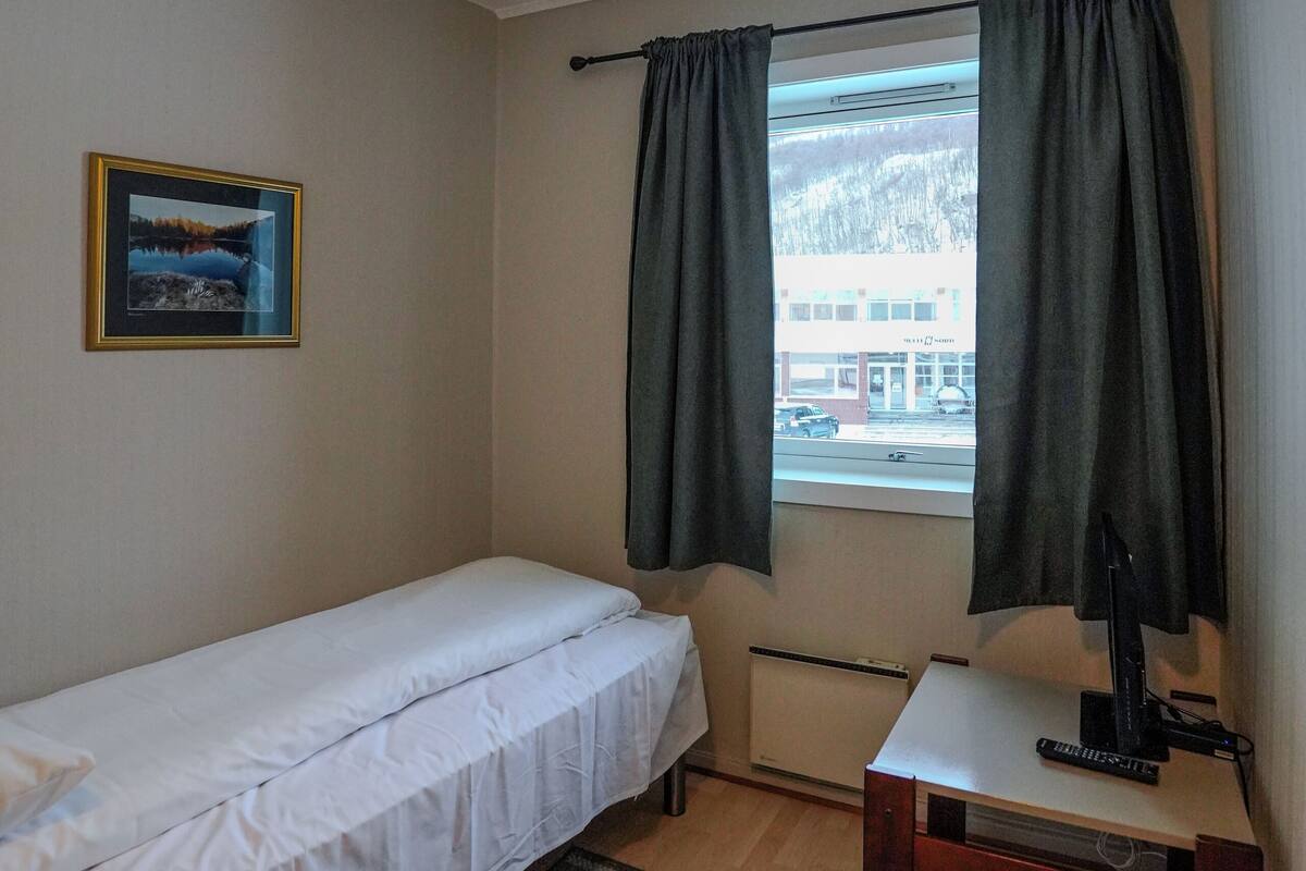 A small, clean room with a single bed, a desk, a chair, a window with curtains, and a framed picture on the wall.