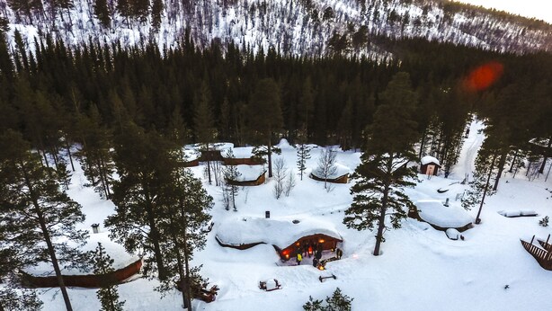 A snow-covered lodge surrounded by trees, with a wooden deck and a small building with a snow-covered roof. - Reisafjord Hotel (Nordreisa)