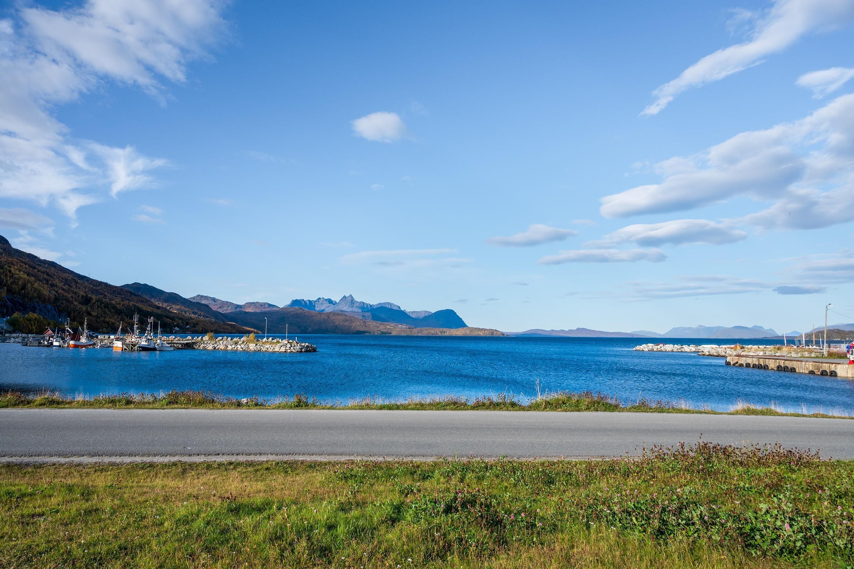 A coastal road with a clear view of a harbor, boats, and mountains in the background.
