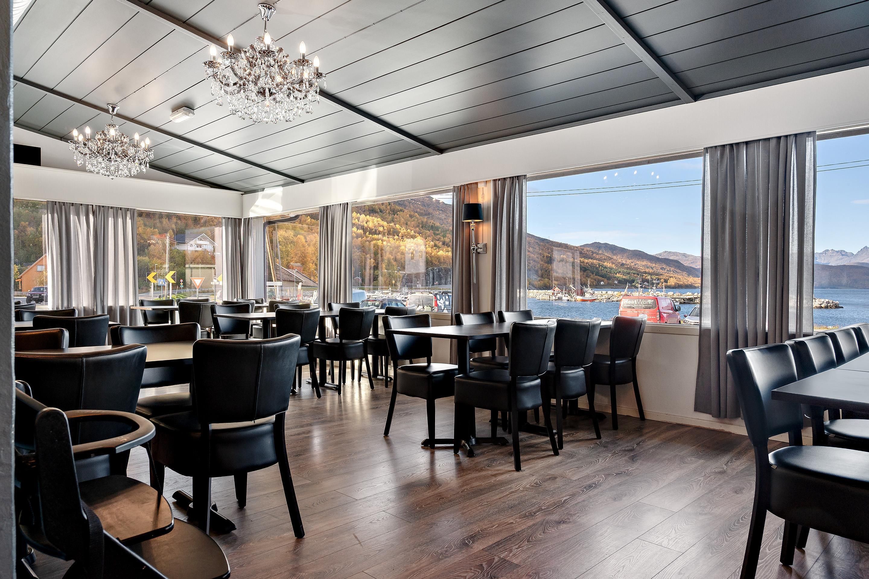 Ein Restaurant mit Blick auf ein Gewässer und Berge, ausgestattet mit Holzböden, schwarzen Stühlen und einer Kristallleuchter.