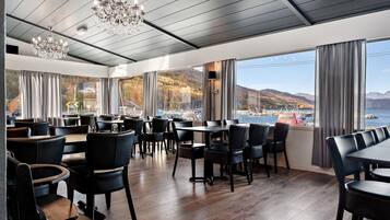 Ein Restaurant mit Blick auf ein Gewässer und Berge, ausgestattet mit Holzböden, schwarzen Stühlen und einer Kristallleuchter.