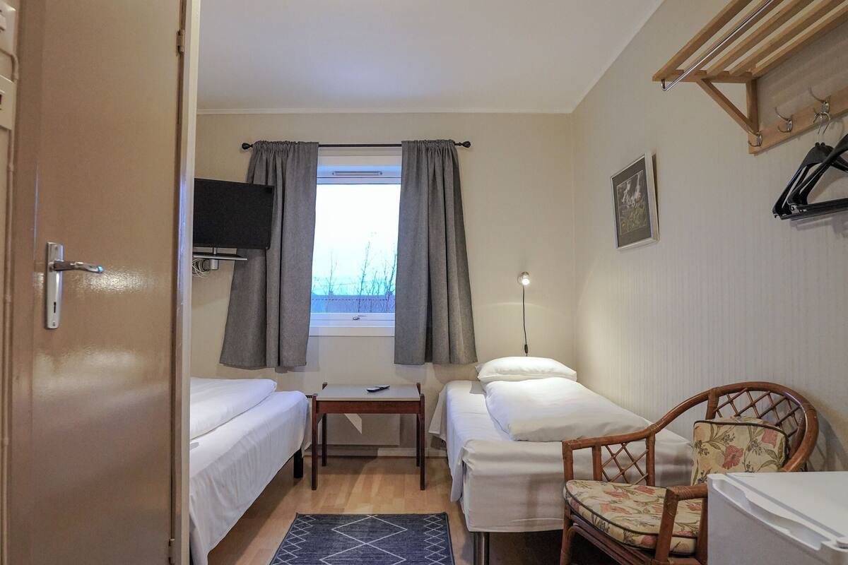 A small, well-lit room with two single beds, a desk, a chair, a radiator, and a window with curtains.