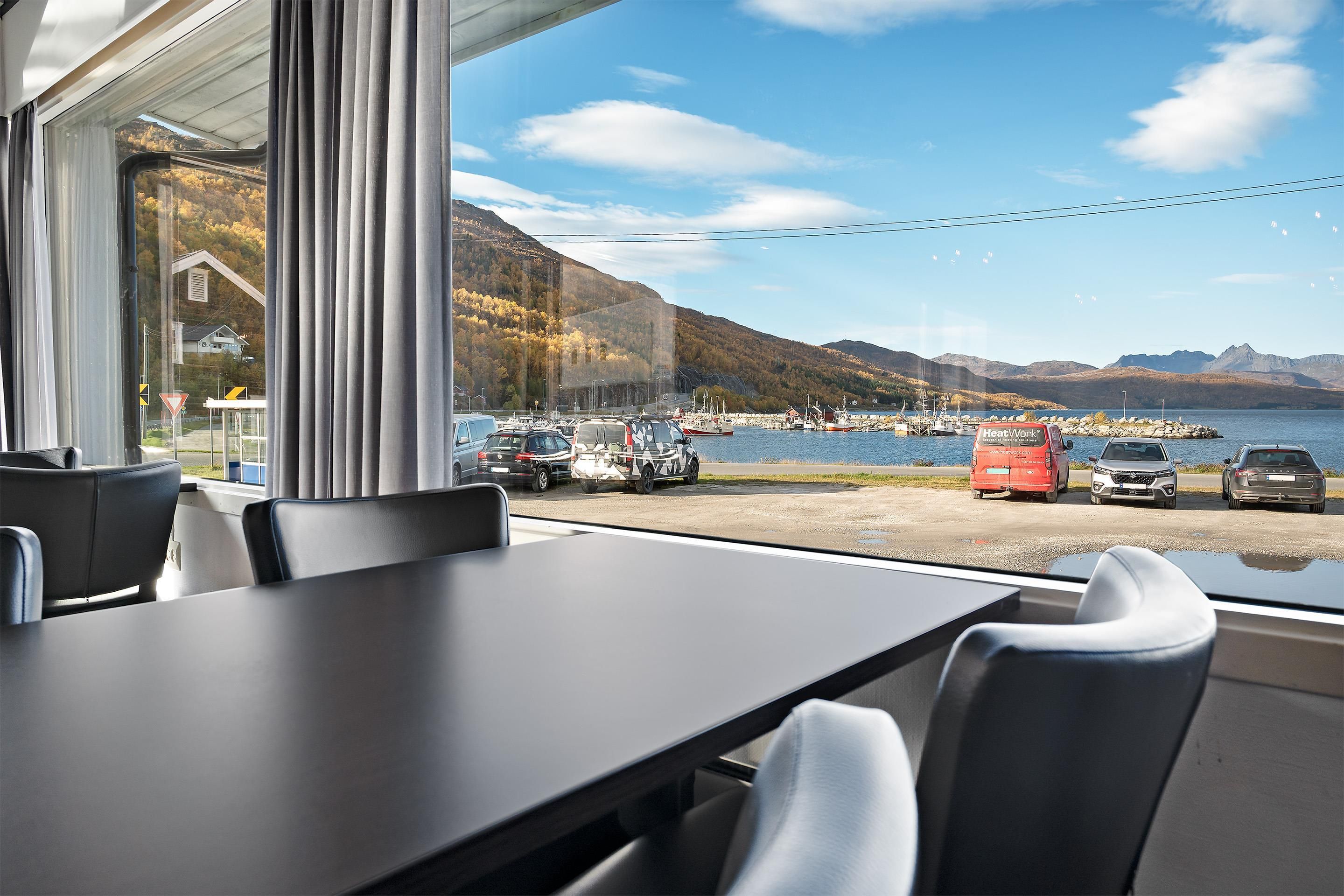 Vue depuis l’intérieur d’un bâtiment, où l’on aperçoit une table et des chaises, donnant sur un port avec des bateaux et un paysage montagneux.