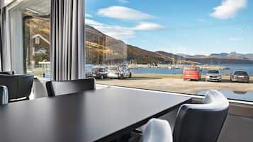 Vue depuis l’intérieur d’un bâtiment, où l’on aperçoit une table et des chaises, donnant sur un port avec des bateaux et un paysage montagneux.