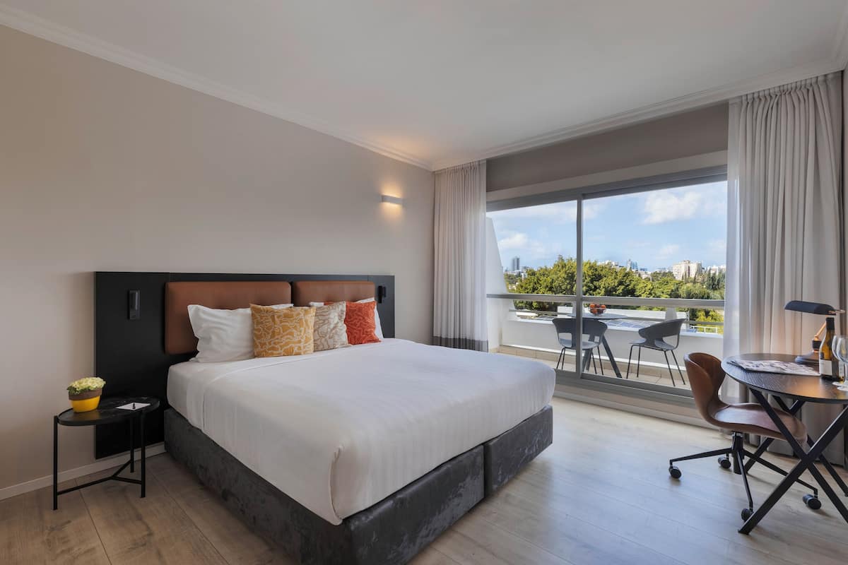 A modern hotel room with a large bed, a desk, and a balcony with a city view.