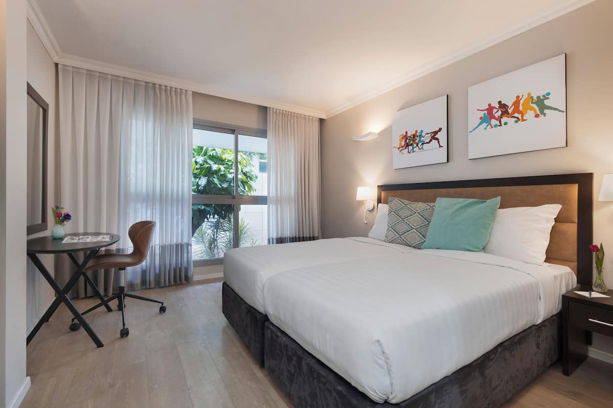A hotel room with a large bed, a desk with a chair, a window with curtains, and two wall-mounted artworks.