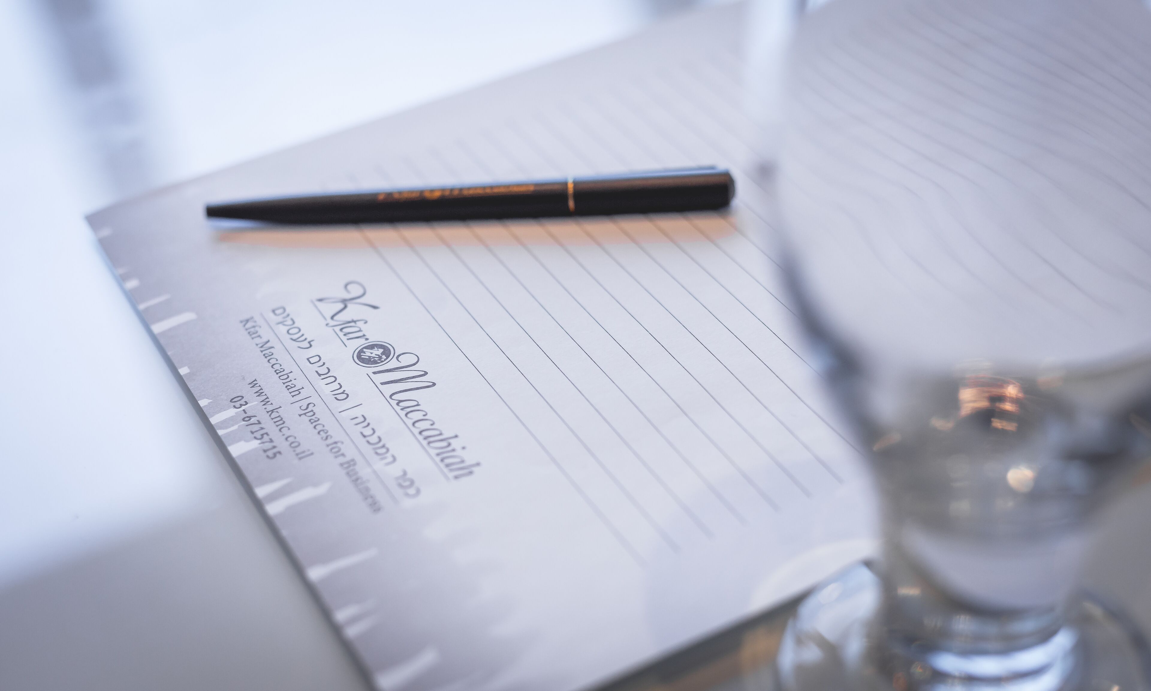 Un cuaderno rayado con una pluma encima, un vaso de agua y un fondo desenfocado.