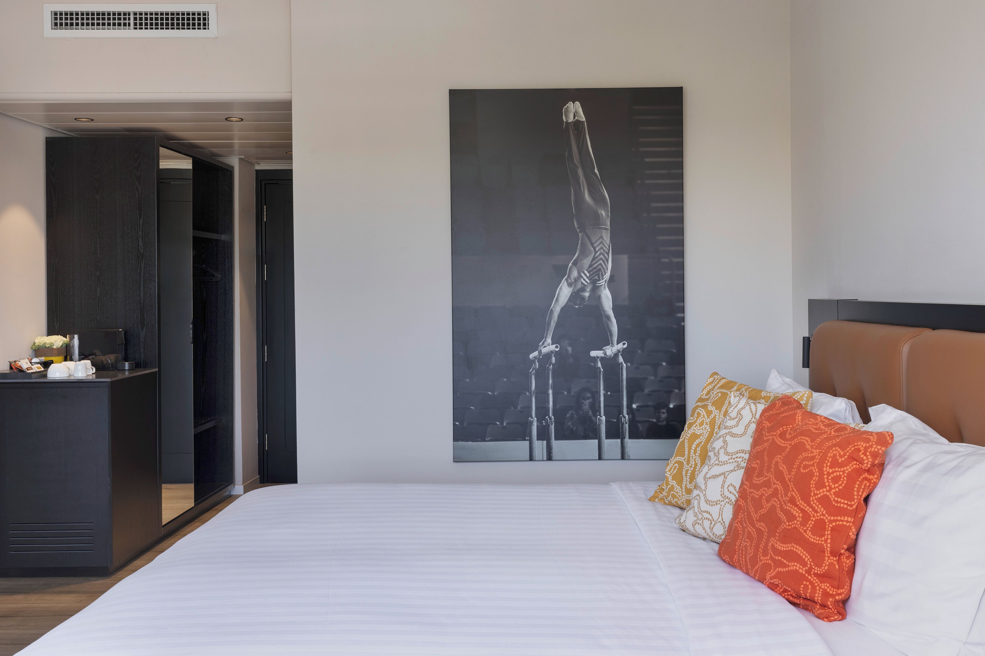 A bedroom with a bed, a nightstand, and a large painting of a gymnast.