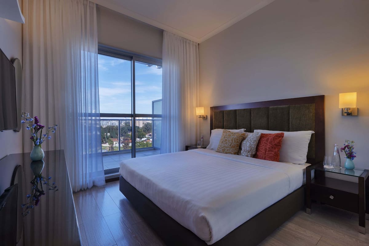 A hotel room with a large bed, a nightstand, a desk, and a view of the city through a large window.