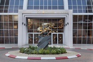 L’entrée d’un hôtel se distingue par une grande façade en verre, une sculpture centrale et un espace paysager agrémenté de plantes.