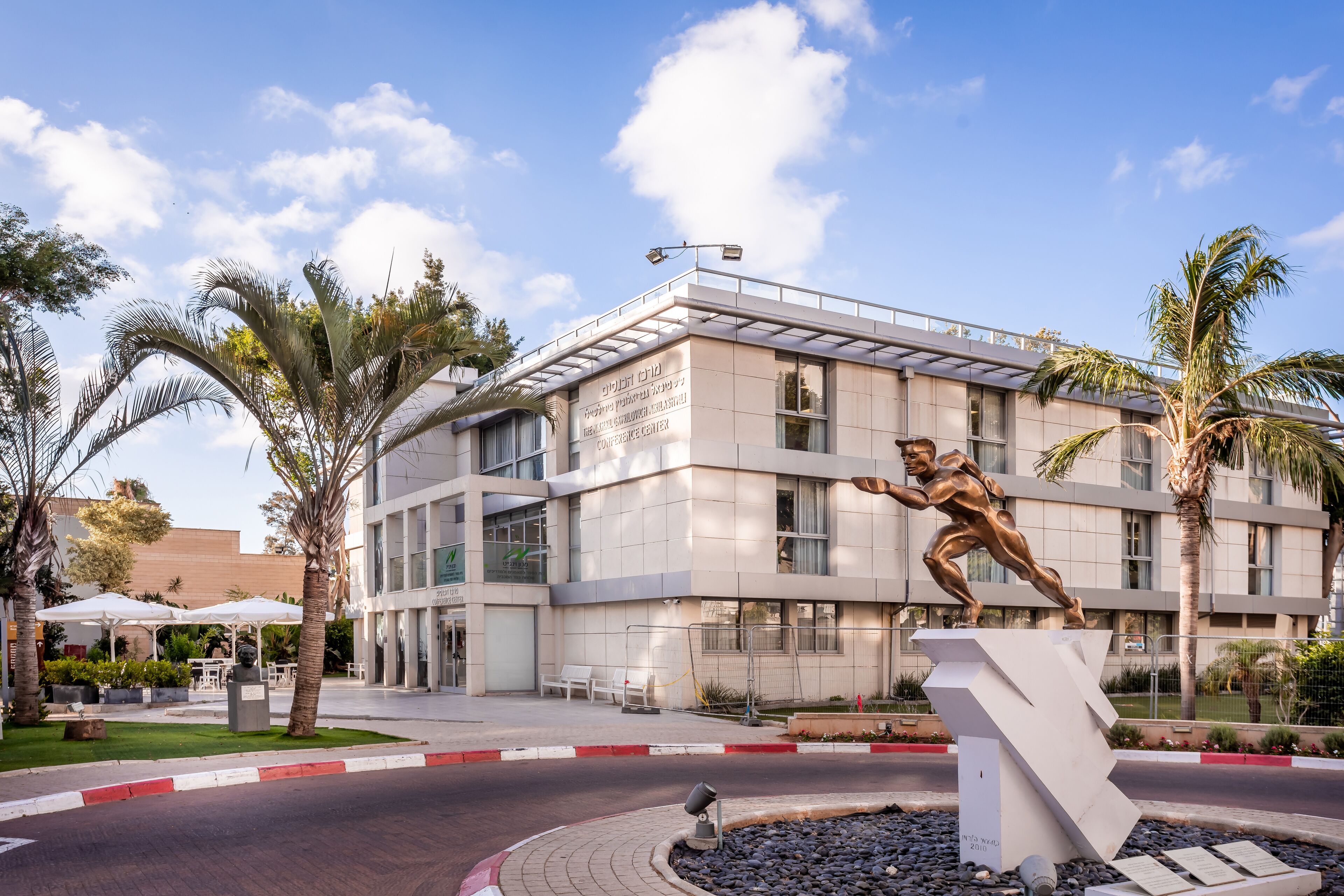 A modern building with a statue of a man in motion, surrounded by palm trees and outdoor seating.