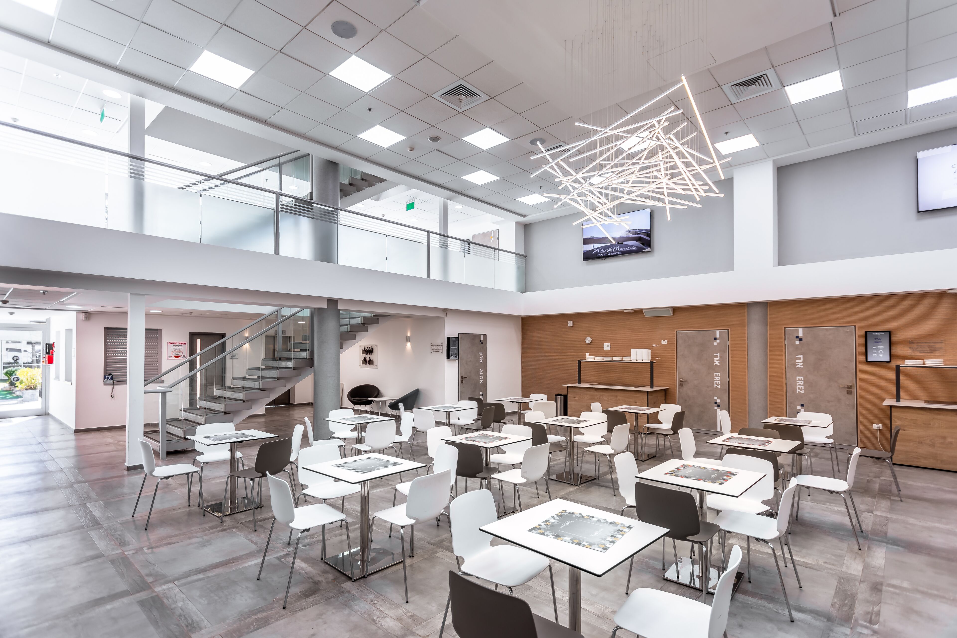 A spacious, modern indoor area with multiple white tables and chairs, a staircase, and a large ceiling light fixture.
