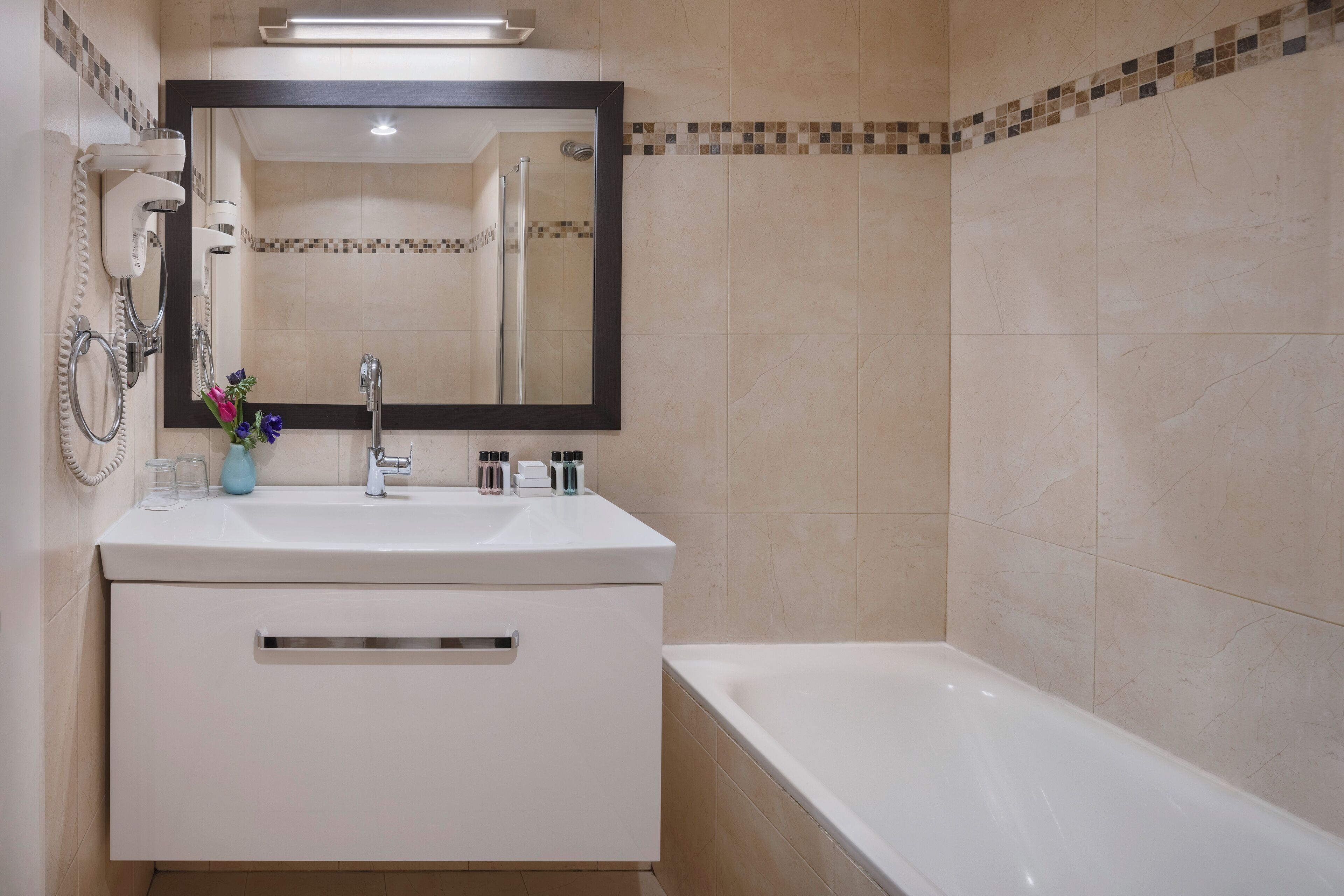 A bathroom with a white vanity, a large mirror, a bathtub, and a tiled wall.