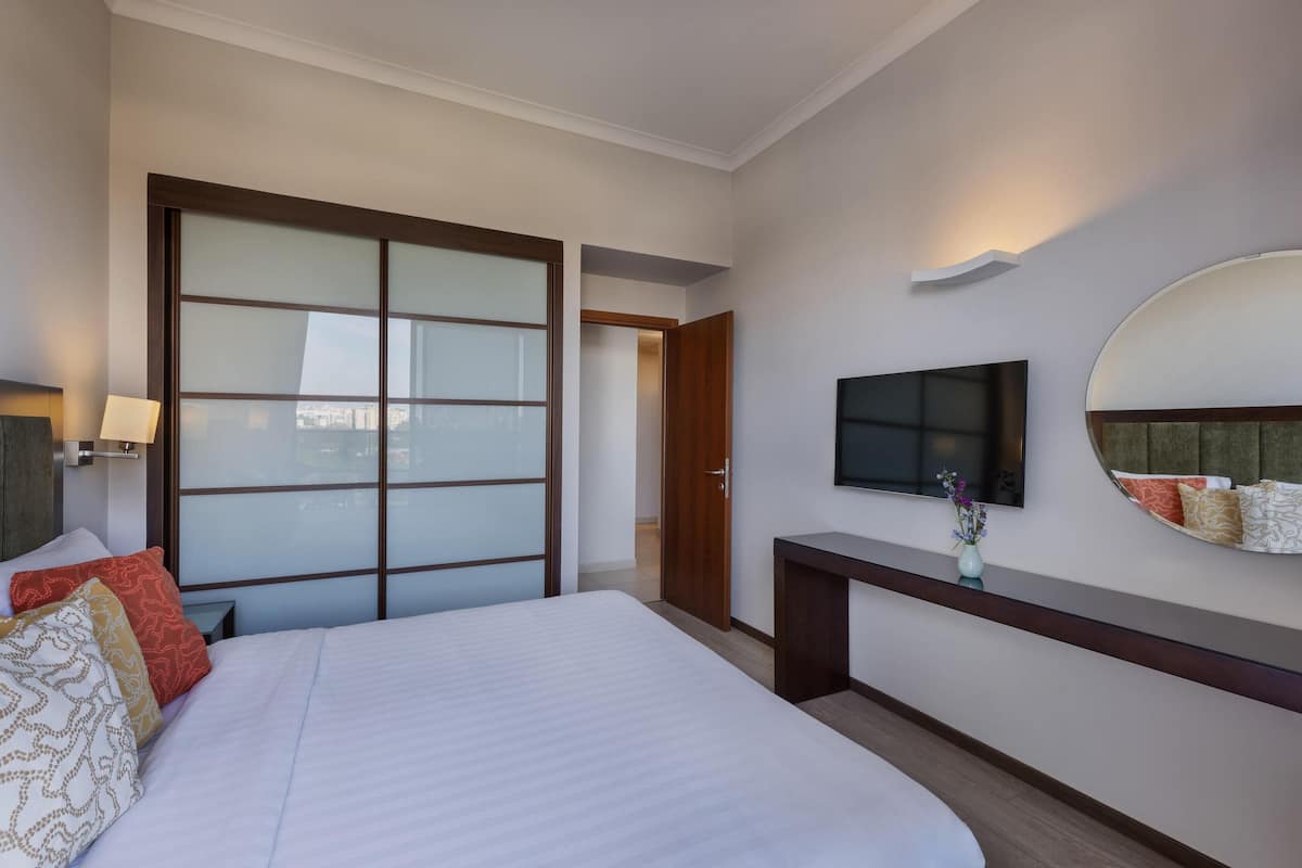 A hotel room with a large bed, a TV mounted on the wall, a wooden desk, and a sliding door leading to another room.