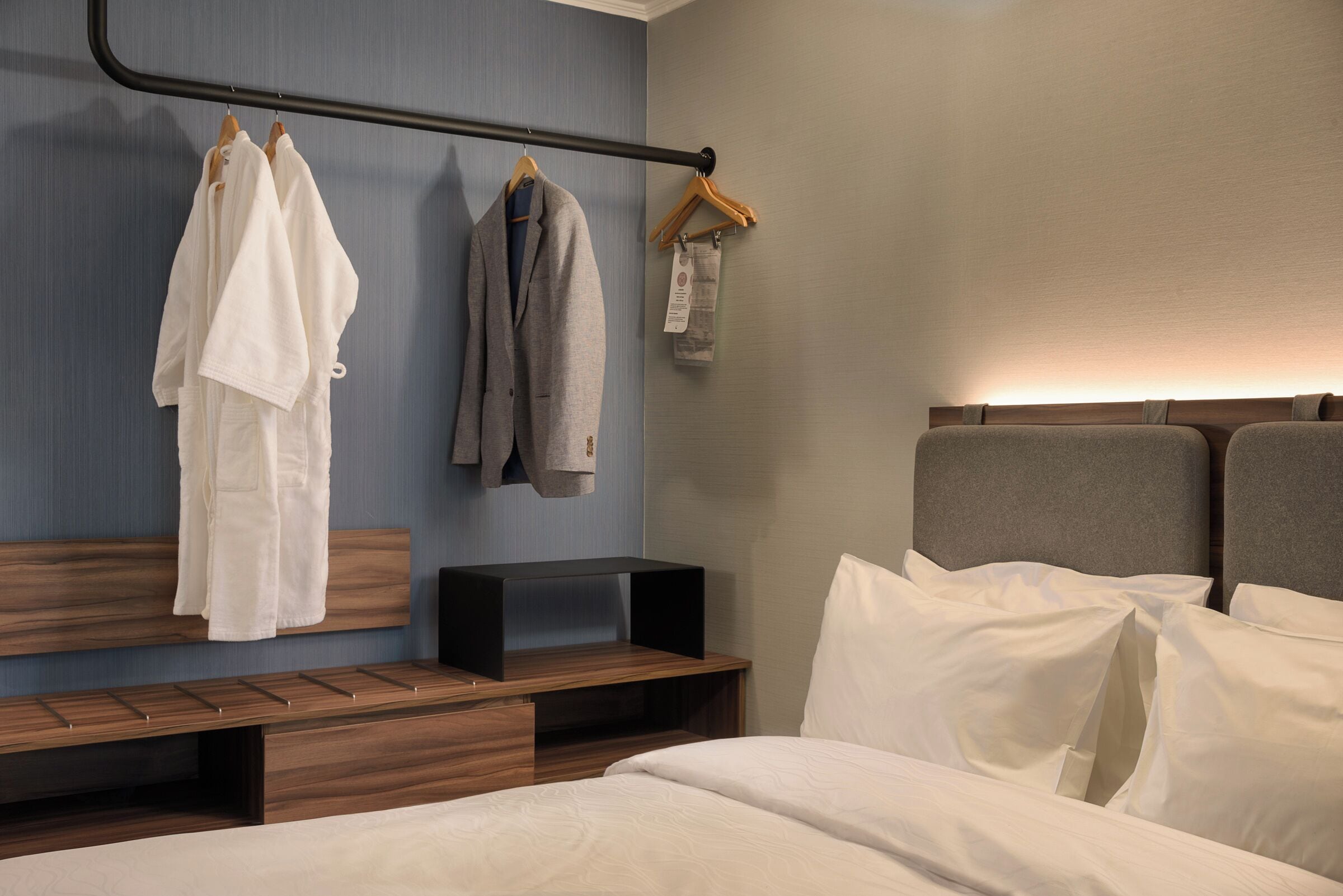 Une chambre d’hôtel comprenant un lit, un banc, une tête de lit en bois et un portant à vêtements avec deux vêtements suspendus.
