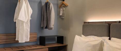 Una habitación de hotel con cama, un banco, cabecero de madera y perchero con dos prendas.