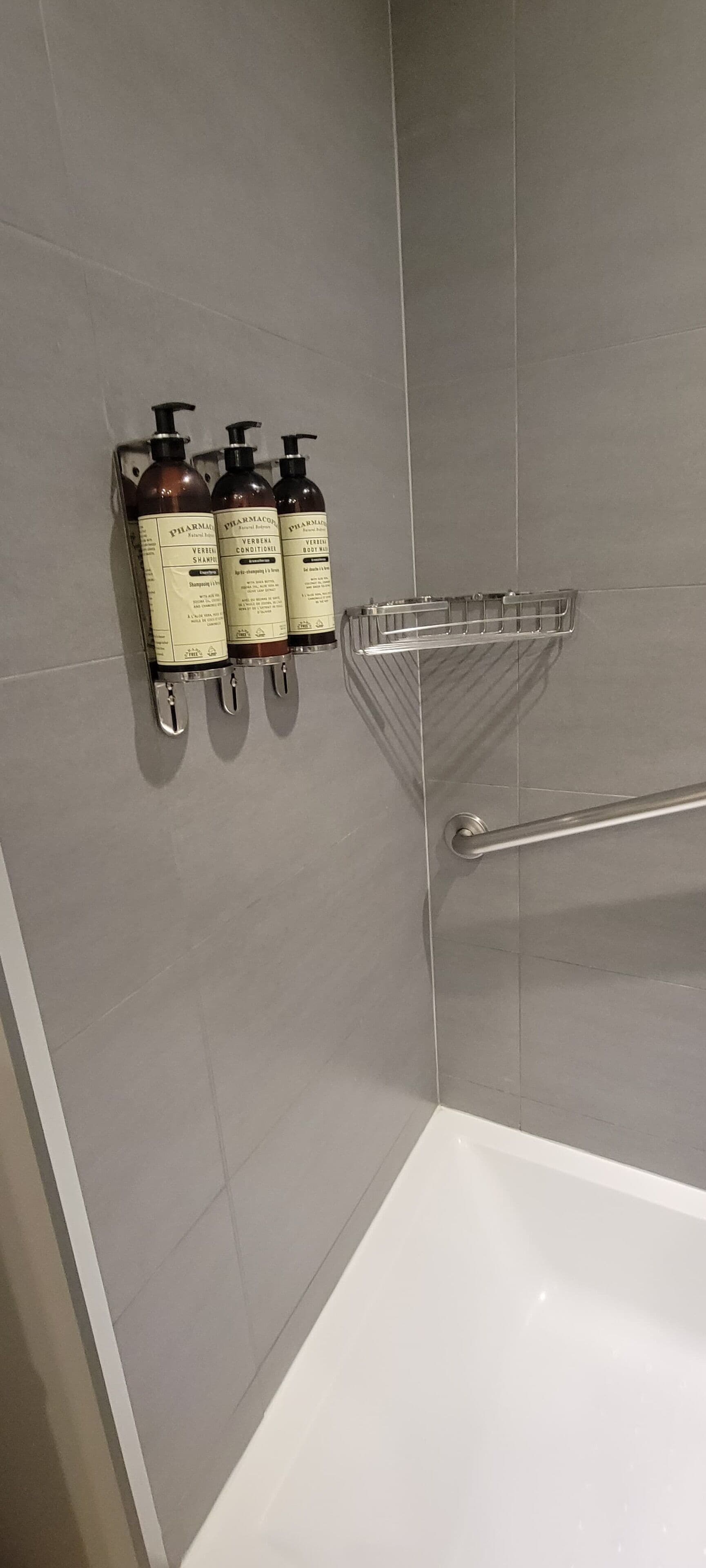 A bathroom with a shower area, a white bathtub, and three bottles of shampoo and conditioner on a wall-mounted rack.
